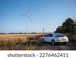 EV car on country ground road near windmills producing renewable energy. Electric automobile on unoccupied site illuminated by sunlight