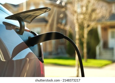 EV electric car plugged charging at residential home under sunset light against blurred street and house around background. Close up.