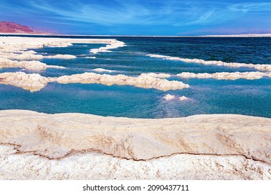 The evaporated salt forms bizarre patterns on the surface of the water. Cirrus clouds fly in the blue sky. Windy spring day. The Israeli coast of the Dead Sea