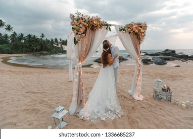 Evening beach with wedding ceremony. Beautiful evening romantic decor and a couple of young newlyweds kissing under the arch. Romantic rustic style. Lamps lights in the evening