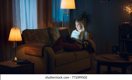 In The Evening Beautiful Young Woman Lies On The Couch With A Laptop On Her Knees. She Types And Has Fun. Room Looks Warm And Cozy.