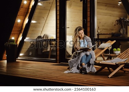As evening falls, a woman wraps herself in a plaid and reads a book on the terrace of log cabin