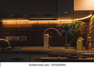Evening life in a country house in the kitchen with a pleasant warm lighting. Modern interior compact kitchen. LED strip with base. Portable drinking water filter with replaceable cartridges.