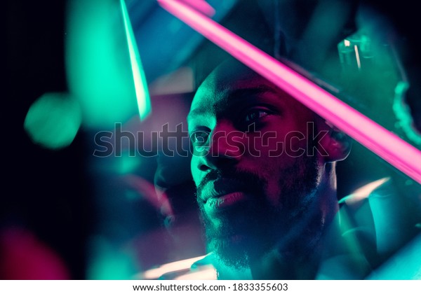 Evening mood. Cinematic portrait of stylish\
young man in neon lighted room. Bright neoned colors.\
African-american model, musician indoors. Youth culture in party,\
festival style and music\
concept.