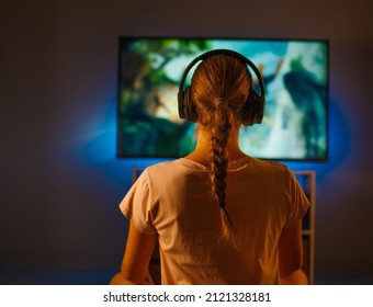 Evening, Night Time. There Is A Blue Neon Light In The Room. A Young Blonde Girl With Headphones Watches TV. Rest, Relaxation, Watching Your Favorite Films And TV Shows.