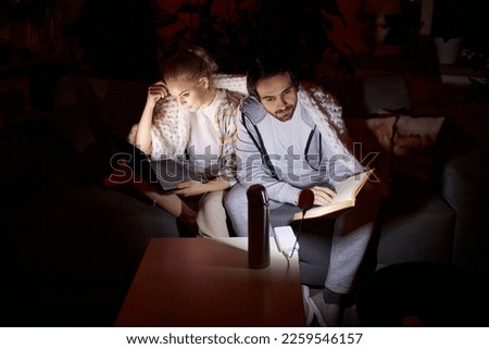 Evening routine. Young couple sitting on sofa, working on laptop and reading book at home in living room. No electricity, blackout. Concept of power outage, family gathering