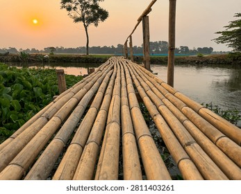 An evening scene featuring a sunset that casts a warm glow over the surrounding landscape. A traditional bamboo bridge spans the river; the structure is situated amidst rice fields.