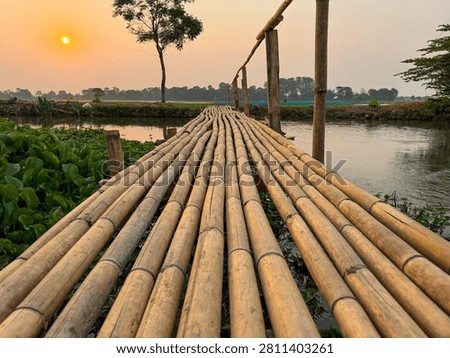 An evening scene featuring a sunset that casts a warm glow over the surrounding landscape. A traditional bamboo bridge spans the river; the structure is situated amidst rice fields.