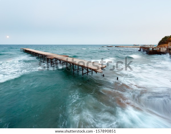 Evening sea surf view, ruined pier\
and Moon in sky (Black Sea, Bulgaria). Long-time\
exposition