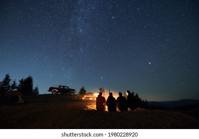 Evening starry sky over mountain valley with car and hikers near campfire. Group of travelers sitting near bonfire under majestic blue sky with stars. Concept of night camping, hiking and travelling. - Shutterstock ID 1980228920