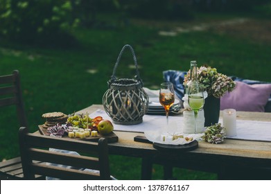 Evening Summer Garden Party With Wine, Cheese And Fruits On Wooden Table Decorated With Candles And Flowers