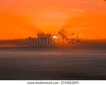 Evening sun drops behind a low cloud bank over the Atlantic off Madeira, casting rays and a glowing silver path on calm slate blue water from a coastal viewpoint