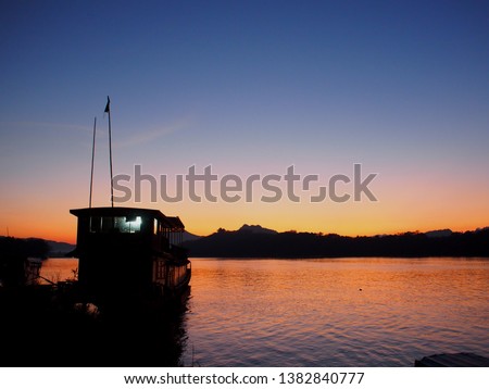 evening sun at horizon with silhouette large wooden house boats on dark water river bank of MEKONG river in LAOS under for unseen natural adventure vacation tour advertisement