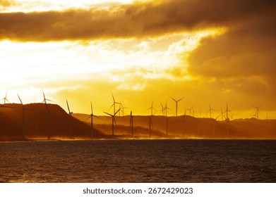 Evening sun pierces dramatic clouds over coastal wind turbines, highlighting clean energy and tranquil ocean reflections