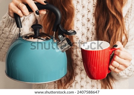 Evening tea making background. Boiling water for hot tea. Woman holding blue hot water kettle. Pouring water into red ceramic mug. Coffee preparation. Gas kettle holding. Girl in woolen sweater.