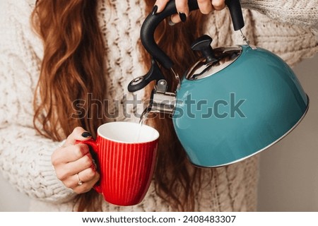 Evening tea making background. Boiling water for hot tea. Woman holding blue hot water kettle. Pouring water into red ceramic mug. Coffee preparation. Gas kettle holding. Girl in woolen sweater.