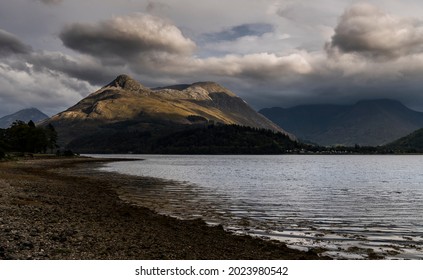 Evening view across bright water of Loch Leven to the sunlit Pap of Glencoe with the mountains at the western end of Glencoe in shadow under low cloud.