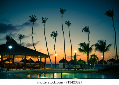 Evening View Of Tropical Resort With Tall Palm Trees, Swimming Pool, Palapa Huts, Beach Bar, Ocean And Sunset.