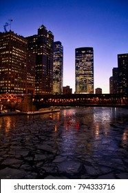 Evening view in winter of elevated train passing through an illuminated Chicago Loop, reflected on a frozen river.