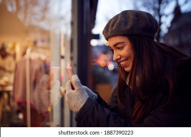 Evening View Of Woman Window Shopping Taking Picture Of Display In Fashion Store On Mobile Phone