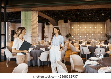 event coordinator with clipboard showing modern banquet hall with decorated tables to young woman