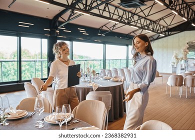 event manager with clipboard showing table with banquet setting to pleased woman in wedding venue