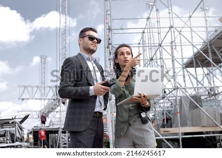 
Event managers discuss stage construction. Installation of stage equipment and preparing for a live concert open air. Event manager portrait. Summer music city festival. Teamwork.