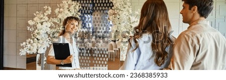 event organizer with clipboard smiling near couple in banquet hall with white floral decor, banner