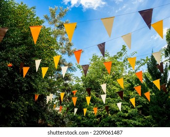 Event Outdoor Party Ornamented With Orange, Yellow, Brown, And White Color Tone Flags In Green Garden On Blue Sky Background. Colorful Triangular Flags Hanging Decorated To Celebrate Halloween Party.