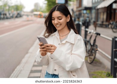 Everyday commute. Cheerful young woman student using the app on her phone while waiting for the public transport - Powered by Shutterstock