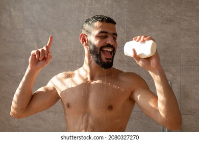 Everyday Hygiene And Relaxation. Positive young bearded man singing taking shower, holding shampoo bottle like microphone standing under falling water drops in modern bathroom at home, pointing finger