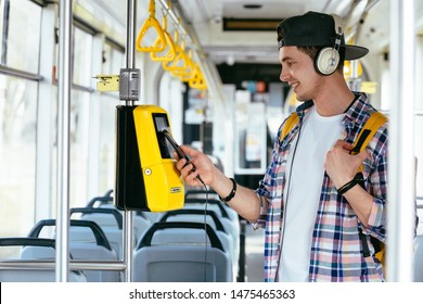 Everyday life and commuting to work or study by bus tram concept. Handsome student man with headphones listening music and paying transport ticket with mobile phone.