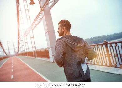 Everyday routine. Back view of young handsome man in sports clothing jogging on the bridge in the morning