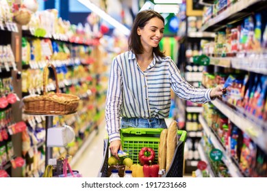 Everyday Shopping Concept. Smiling Young Housewife Pushing Trolley Cart Between Aisles In Grocery Store. Positive Woman Buying Food, Taking Products From Shelf. Consumerism And Lifestyle - Shutterstock ID 1917231668