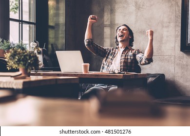Everyday winner. Cheerful young man in casual wear keeping arms raised and looking happy while sitting at the desk in office 