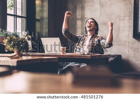 Everyday winner. Cheerful young man in casual wear keeping arms raised and looking happy while sitting at the desk in office 