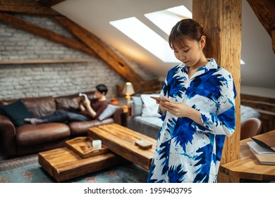 Everyone Has Own Entertainment. Asian Female Using Cellphone While Her Boyfriend Lying On Sofa And Reading Book