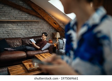 Everyone Has Own Entertainment. Asian Female Using Cellphone While Her Boyfriend Lying On Sofa And Reading Book