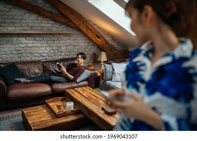 Everyone Has Own Entertainment. Japanese Female Using Cellphone While Her Boyfriend Lying On Sofa And Reading Book