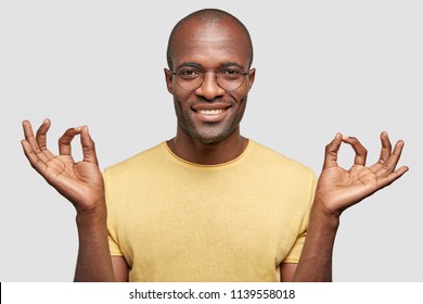 Everything is okay! Satisfied bald dark skinned male with positive smile, gestures indoor, dressed in casual yellow t shirt, isolated over white background. People and body language concept.