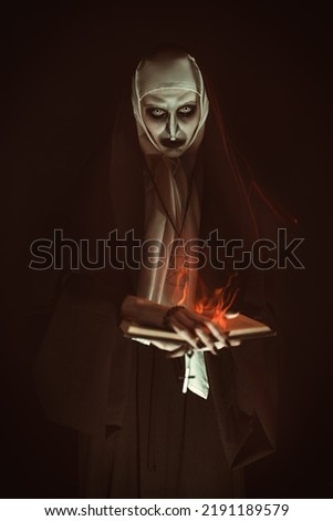 An evil cursed nun with a burning bible and a cross in her hands looks at the camera on a black background. Halloween. Horrors. 