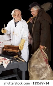 Evil Doctor Brandishing Cleaver Exchanges Glances With Grave Robber Over Bloody Corpse. Stage Effect, Isolated On Black Background, Spot Lighting.