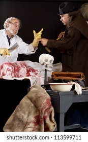 Evil Doctor Interacts With Grave Robber Over Bloody Corpse. Stage Effect, Isolated On Black Background, Spot Lighting.