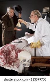 Evil Doctor Reaches Over Bloody Corpse And Pays Grave Robber, Who Tips Hat. Stage Effect With Dark Background, Spot Lighting.