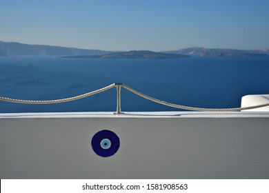 Evil Eye Charm On A Whitewashed Wall With A Rope Railing Overlooking The Caldera In Oia Santorini Island, Cyclades Greece. 