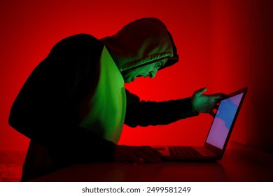 Evil hacker in front of his laptop 
An individual wearing a green and black hoodie sits in front of a laptop, visibly frustrated. The room is illuminated with a striking red light. - Powered by Shutterstock