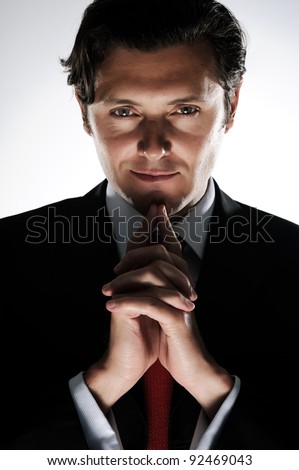 Evil looking businessman clasps his hands under his chin, in dramatic lighting
