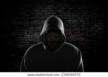An evil looking man with a hidden face and wearing a hoodie stands intimidatingly against an ally wall stalking his next victim.

