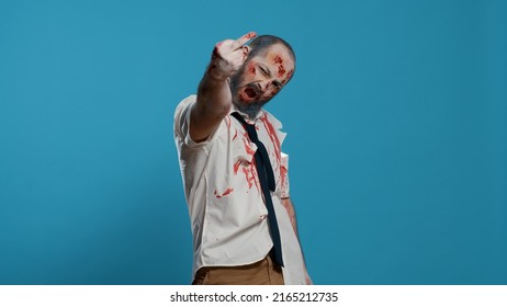 Evil Looking Scary Mindless Zombie Being Rude By Showing Middle Finger To Camera. Sinister Frightening Brain-dead Walking Corpse Making Rude Finger Gesture On Blue Background.