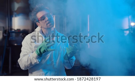 Evil professor pushing remote controller button in large industrial laboratory. Portrait of crazy male scientist in lab coat, protective gloves and goggles releasing blue smoke and laughing